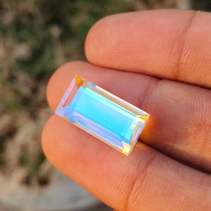 May include: A rectangular, iridescent gemstone with a light blue centre and a rainbow-coloured edge. The gemstone is held in a person's hand, with a blurred green background.