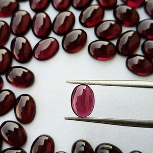 May include: A collection of deep red, oval-shaped cabochon gemstones with a smooth, polished finish. One single oval gemstone is held between the metal tips of a pair of tweezers, showcasing its translucent, dark crimson color and rounded surface.