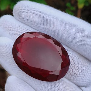 Può includere: Una gemma sfaccettata, di forma ovale, di colore rosso intenso. La gemma è tenuta in una mano guantata di bianco, mostrando la sua superficie lucida e i suoi intricati tagli. Il colore della gemma è un rosso scuro e ricco.