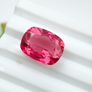 May include: A faceted, cushion-cut gemstone in a vibrant shade of pink. The gem is set against a white, ridged surface, highlighting its color and clarity. The gemstone's facets catch the light, creating a sparkling effect. A beautiful piece for jewelry making.