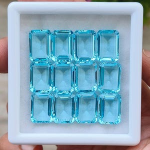 May include: A white square box containing twelve rectangular, light blue gemstones. The gemstones are arranged in three rows of four. The gems have a faceted cut, reflecting light. The box is held by a person's hand.