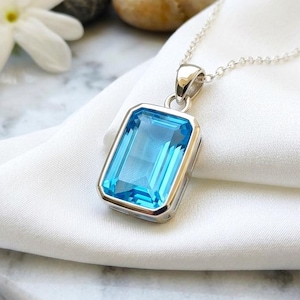Peut inclure: Un pendentif rectangulaire en pierre bleue taille émeraude, serti dans une monture en métal argenté poli et suspendu à une fine chaîne en argent. La pierre bleue vive présente des bords facettés qui captent la lumière avec éclat.