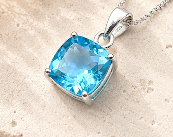 May include: A silver pendant necklace featuring a cushion-cut, bright blue gemstone set in a four-prong setting. The metal bail is stamped with 925, indicating sterling silver. The pendant hangs from a delicate silver chain with a polished finish.