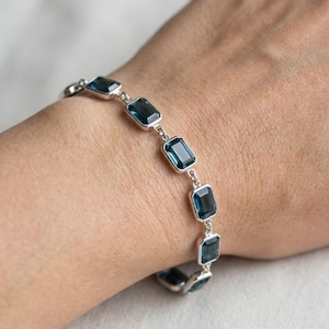May include: A silver bracelet featuring rectangular, deep blue gemstones. The bracelet is composed of multiple links, each holding a single gem. The gems are set in a silver metal, creating a contrast between the dark blue stones and the bright metal.