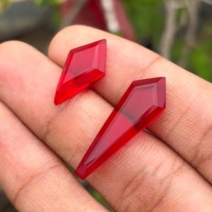May include: Two faceted red gemstone pieces with a translucent appearance. One piece is a small diamond shape, and the other is a longer, tapered, coffin-like geometric shape. Both items feature sharp, clean edges and a smooth, polished surface finish.
