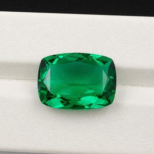 May include: A large, emerald-green gemstone with a cushion cut. The gem is displayed on a white surface, showcasing its vibrant color and faceted design. The gemstone's polished surfaces reflect light, creating a sparkling effect.