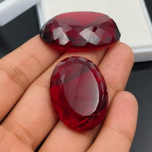 Pode incluir: Duas pedras preciosas vermelhas escuras, ovais e facetadas. Uma gema é exibida em uma mão, enquanto a outra repousa perto de uma caixa de exibição branca. As gemas têm um corte brilhante, refletindo a luz.