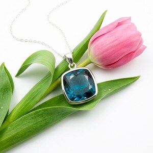 May include: A silver necklace with a large, square, teal-colored pendant. The pendant is set in a silver frame and hangs from a delicate chain. The necklace rests on a green leaf and a pink tulip.