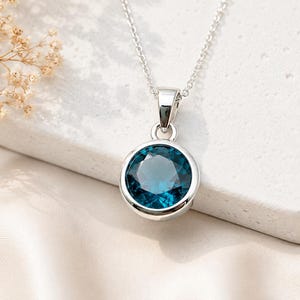 Può includere: Una collana con pendente in argento con una gemma rotonda color teal. La gemma è incastonata in una lunetta d'argento e pende da una delicata catena d'argento. La collana è esposta su una superficie bianca con uno sfondo morbido e neutro.