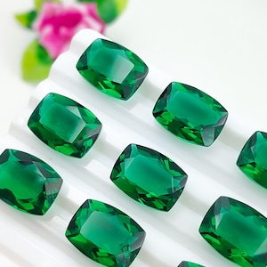 Pode incluir: Uma coleção de pedras preciosas verde-esmeralda vibrantes, cada uma com um corte almofada facetado. Estas pedras translúcidas de formato retangular estão dispostas numa grelha sobre um tabuleiro branco, realçando a sua cor profunda e clareza.