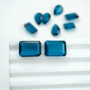 Pode incluir: Duas pedras preciosas azul-profundo com corte esmeralda estão posicionadas lado a lado em primeiro plano. No fundo desfocado, várias outras pedras preciosas soltas de formas variadas, incluindo gota e retangular, estão espalhadas.