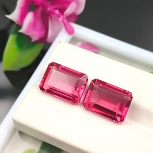 May include: Two rectangular, emerald-cut gemstones with a vibrant, deep pink hue. These faceted stones exhibit high clarity and transparency, reflecting light across their polished surfaces. They are positioned side-by-side on a white display stand.