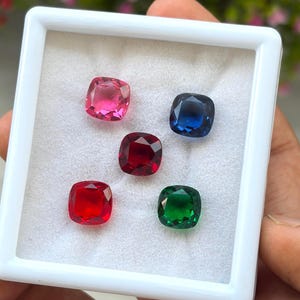 Può includere: Una scatola espositiva quadrata bianca contenente cinque pietre preziose sfaccettate a taglio cuscino disposte a grappolo. La collezione comprende una pietra rosa, una blu scuro, una rosso intenso, una rosso brillante e una verde smeraldo su velluto.