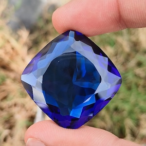 May include: A large, square-cut, faceted gemstone with a deep blue hue. The gem is held between two fingers, showcasing its intricate internal structure and reflective surfaces. The background is blurred, emphasizing the gem's clarity and color.