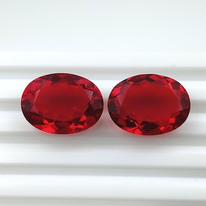 May include: Two oval-shaped, faceted gemstones with a deep red color. The gems are displayed against a white background with horizontal lines. The facets create a sparkling effect, highlighting the rich color and clarity of the stones. Ideal for jewelry making.