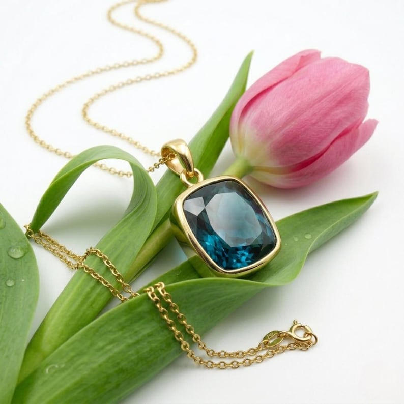 May include: A gold necklace with a large, square, teal-colored gemstone pendant. The pendant is set in a gold bezel. The necklace rests on a green leaf, next to a pink tulip. The chain is delicate and the clasp is visible.