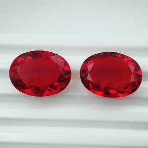 May include: Two oval-cut, faceted gemstones with a deep red color. The gems are displayed on a white surface, showcasing their brilliance and clarity. The shape and color suggest they could be rubies or similar precious stones.