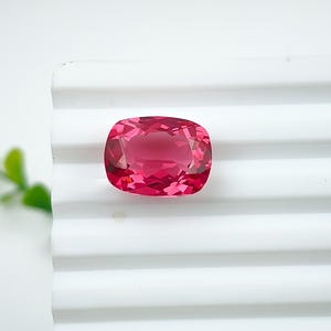 May include: A cushion-cut, faceted gemstone in a vibrant pink hue. The gem is set against a white, ridged surface, showcasing its clarity and brilliance. The gemstone's shape is rectangular with rounded corners, reflecting light beautifully.
