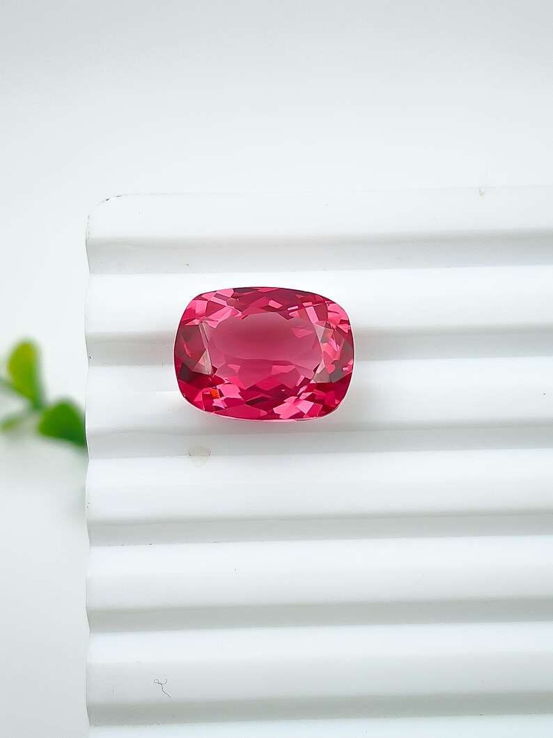 May include: A cushion-cut, faceted gemstone in a vibrant pink hue. The gem is set against a white, ridged surface, showcasing its clarity and brilliance. The gemstone's shape is rectangular with rounded corners, reflecting light beautifully.