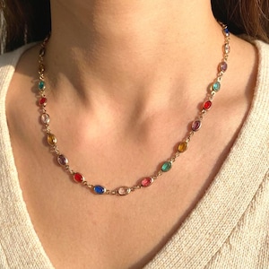 Puede incluir: Un collar de cadena en tono dorado que presenta una serie de pequeñas piedras preciosas ovaladas y multicolores engastadas en biseles de metal individuales. Las piedras muestran una variedad de tonos vibrantes que incluyen rojo, azul, verde, amarillo y morado, dispuestos en un patrón repetitivo a lo largo de la delicada cadena de eslabones.