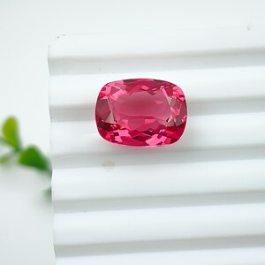 May include: A faceted, cushion-cut gemstone in a vibrant shade of pink. The gem is set against a white, ridged surface, with a soft, diffused light. The gemstone's facets reflect light, creating a sparkling effect.