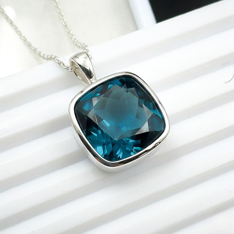 May include: A silver pendant necklace featuring a large, square-cut, teal-colored gemstone. The gemstone is set in a silver bezel and hangs from a delicate silver chain. The pendant has a polished, reflective surface, and the overall design is elegant and modern.