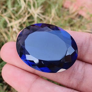 Puede incluir: Una gema grande, facetada, de forma ovalada y color azul profundo. La gema se sostiene en una mano, con un fondo borroso de hierba verde. Las facetas crean un juego de luces y sombras.
