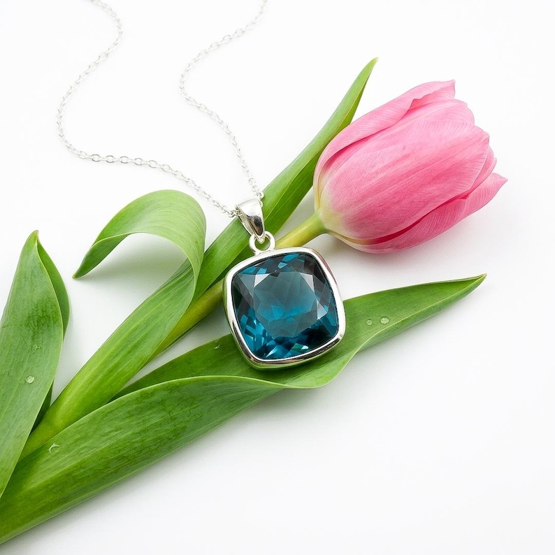 May include: A silver necklace with a large, square, teal-colored pendant. The pendant is set in a silver frame and hangs from a delicate chain. The necklace rests on a green leaf and a pink tulip.