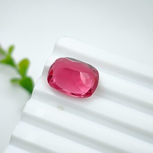 May include: A faceted, rectangular-shaped gemstone in a vibrant shade of pink. The gem is set against a white, ridged surface, with a few green leaves visible in the background. The gem's cut allows light to reflect, creating a sparkling effect.