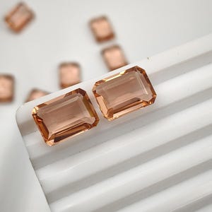 Può includere: Due pietre preziose rettangolari, sfaccettate e color pesca con un aspetto chiaro e trasparente. Le pietre presentano bordi netti e puliti e una superficie lucida, che riflette la luce sulle loro sommità piatte. Sono visibili diverse pietre identiche.