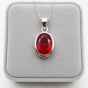 May include: A silver pendant necklace with an oval, faceted red gemstone. The gem is set in a silver bezel and suspended from a delicate silver chain. The necklace is presented on a grey velvet surface.