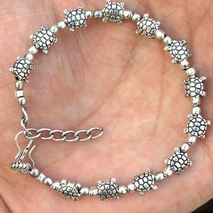 Puede incluir: Una pulsera de tono plateado que presenta una serie de pequeños dijes de tortuga detallados que se alternan con cuentas redondas y lisas. La pulsera incluye una cadena extensora de metal y un cierre de tipo gancho.