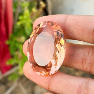 Può includere: Una grande gemma sfaccettata di forma ovale con un aspetto traslucido color pesca pallido. La pietra presenta numerosi tagli geometrici sulla superficie, creando un aspetto riflettente e limpido che cattura la luce da diverse angolazioni.