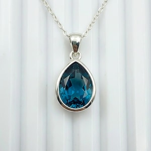 Può includere: Collana in argento con un pendente a forma di goccia. Il pendente presenta una gemma blu-verde intenso sfaccettata, incastonata in una lunetta d'argento, sospesa a una delicata catena d'argento. Sfondo bianco strutturato.