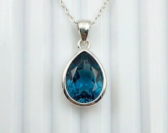 May include: A silver necklace featuring a teardrop-shaped pendant with a faceted, deep teal gemstone. The pendant is set in a silver bezel and hangs from a delicate silver chain. The background is a white, textured surface.