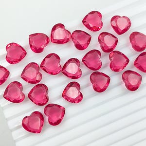 May include: A collection of faceted, heart-shaped, pink gemstones arranged on a white, ridged surface. The gemstones have a glossy, transparent appearance, reflecting light. The hearts vary slightly in size and are scattered across the surface.