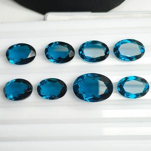 May include: Eight oval-cut, deep blue gemstones arranged in two rows. Each stone features a faceted surface that reflects light, showcasing a consistent dark blue hue across all pieces. The stones are displayed on a white, ridged surface.