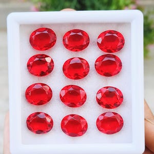 Puede incluir: Una caja de presentación rectangular blanca que contiene doce piedras preciosas rojas de forma ovalada y talladas, dispuestas en una cuadrícula de cuatro por tres. Cada piedra presenta un corte brillante con múltiples superficies reflectantes.