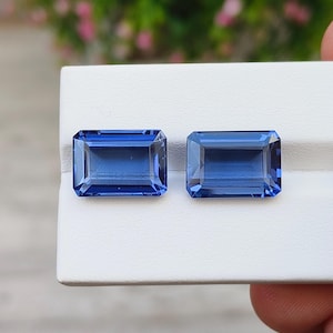 Può includere: Due pietre preziose blu rettangolari e sfaccettate con un colore profondo e saturo e una trasparenza limpida. Le pietre sono posizionate fianco a fianco, mostrando un classico taglio a smeraldo con angoli vivi e molteplici sfaccettature.