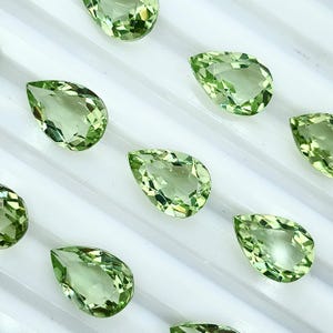 May include: A collection of light green, pear-shaped gemstones with faceted surfaces. The gems are arranged on a white surface, showcasing their clarity and color. The gemstones are likely for jewelry making or collecting.