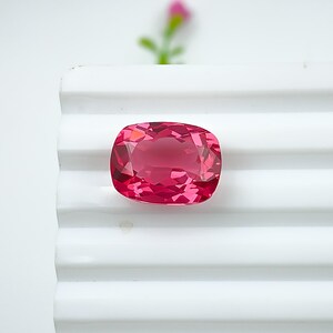 May include: A faceted, cushion-cut, pink gemstone. The gem is a vibrant shade of pink, with multiple facets that reflect light. It is set against a white, ridged surface, with a blurred pink flower in the background. The gemstone is likely for jewelry.