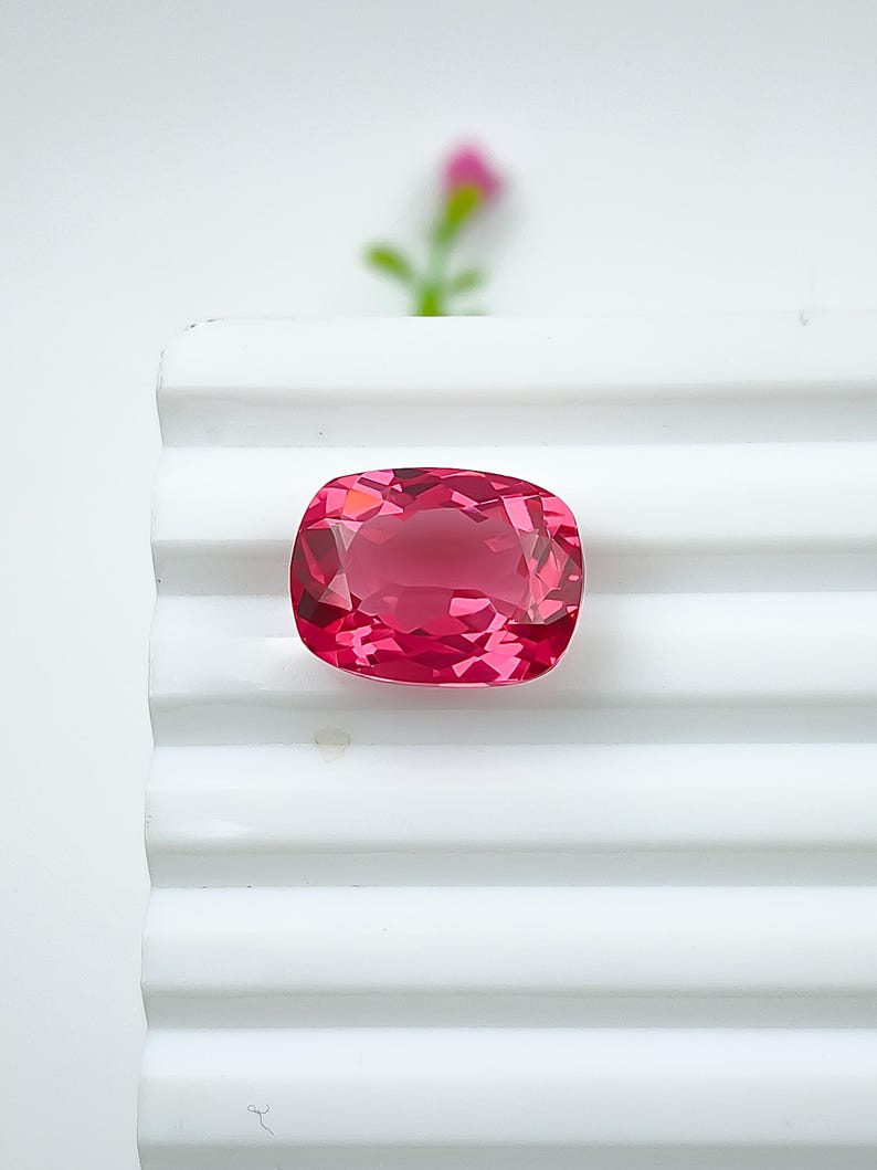 May include: A faceted, cushion-cut, pink gemstone. The gem is a vibrant shade of pink, with multiple facets that reflect light. It is set against a white, ridged surface, with a blurred pink flower in the background. The gemstone is likely for jewelry.