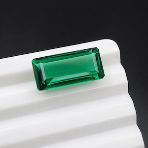 Puede incluir: Una gema rectangular de color verde esmeralda con un corte facetado. La gema está colocada sobre una superficie blanca y acanalada, con un fondo negro. El color vibrante y los cortes precisos de la gema se destacan.