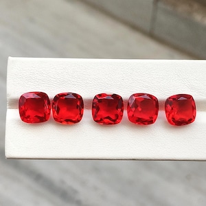 May include: Five vibrant red gemstones with a cushion cut, arranged in a horizontal row. Each stone features multiple facets that reflect light, creating a brilliant appearance. The gems are set against a white, textured display surface.