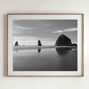 May include: A black and white photograph of Haystack Rock and other rock formations on the Oregon coast. The rocks are reflected in the calm water of the ocean.