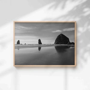 Peut inclure: Photographie encadrée en noir et blanc d'une scène de plage. L'image présente trois grandes formations rocheuses dans l'océan, avec le sable mouillé reflétant le ciel et les nuages. La photographie est dans un cadre en bois.