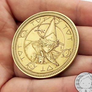 Puede incluir: Una moneda circular de metal dorado con una ilustración en relieve de un personaje con sombrero puntiagudo y gafas, con un dedo levantado. El borde contiene símbolos grabados, incluidos triángulos y formas circulares similares a ojos.