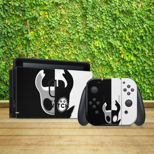 Peut inclure: Une console et une manette Nintendo Switch noires et blanches avec un graphisme d'une créature du jeu vidéo Hollow Knight. La console et la manette sont sur une surface en bois.