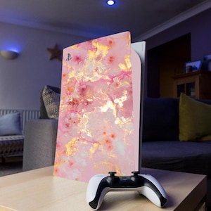 May include: Pink and gold marble design Playstation 5 console skin with the Playstation logo. The console is standing on a table with a black and white Playstation 5 controller.