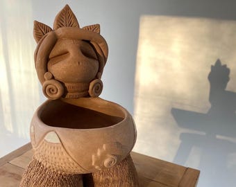 May include: A terracotta planter shaped like a woman with a closed-eye face and a crown of leaves. The planter has a wide, round opening for planting.